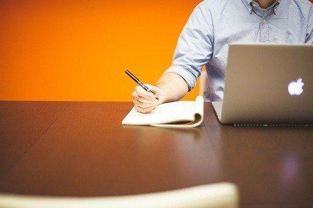 A man is sitting at a table with a laptop and writing in a notebook.